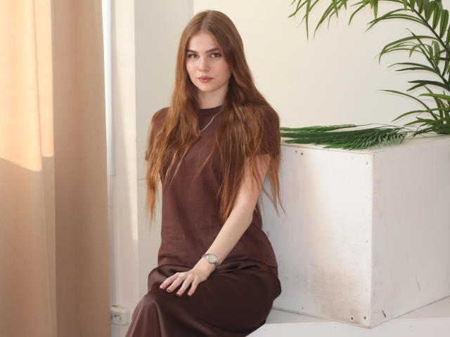 MargoKoli with long brown hair, brown dress, sitting on white box, green plant, watch on wrist, soft light, neutral expression, beige curtain, relaxed pose.