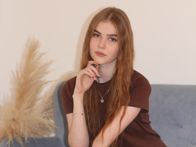 MargoKoli, avec de longs cheveux roux, un t-shirt marron, assise sur un canapé bleu, la main près du menton, regardant la caméra, de l'herbe beige duveteuse sur la gauche, un collier, un léger sourire, décontractée..