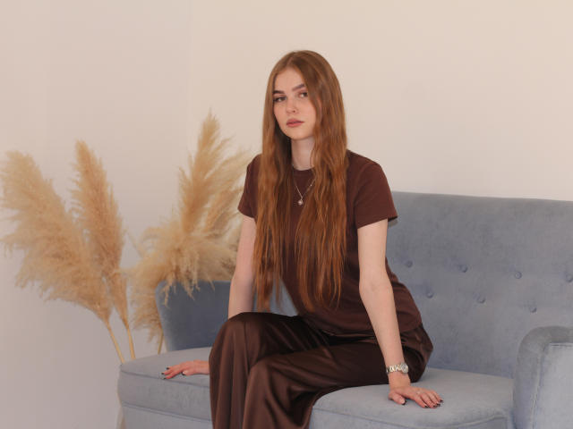 MargoKoli with long red hair, brown dress, and silver watch, sitting on a blue couch, puffy tan grasses behind her, looking serious, hand on couch, relaxed posture.
