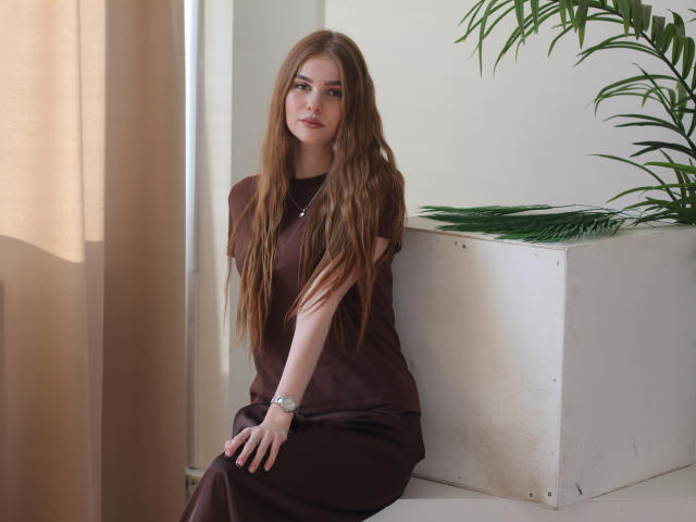 MargoKoli, com cabelos castanhos longos, vestido marrom, sentada em uma caixa branca, relógio no pulso, planta verde à direita, cortina bege à esquerda, luz suave, expressão calma, aparência natural..