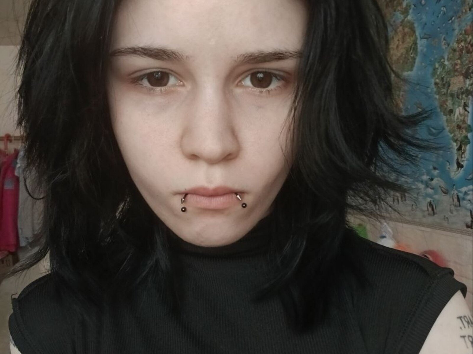 JaneneRandy, avec ses cheveux noirs, sa peau pâle, ses yeux marrons et son pull noir à col roulé, a un piercing au nez et un anneau aux lèvres avec des boules noires. Son expression est neutre et légèrement sérieuse..