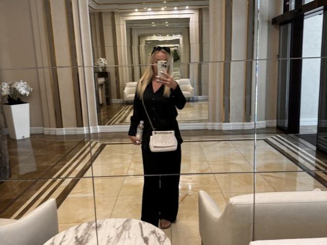 Een mysterieuze vrouw met lang blond haar, gekleed in een zwarte outfit en met een telefoon in haar hand, staat in een gang met spiegels. Ze heeft een klein achterwerk en draagt een witte, gewatteerde crossbodytas..