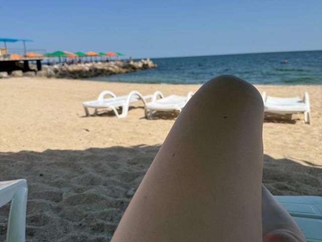 MysteryLaddy's leg in the foreground, pale skin, showing a slight tan line, with a sandy beach, blue ocean, and white lounge chairs in the background.