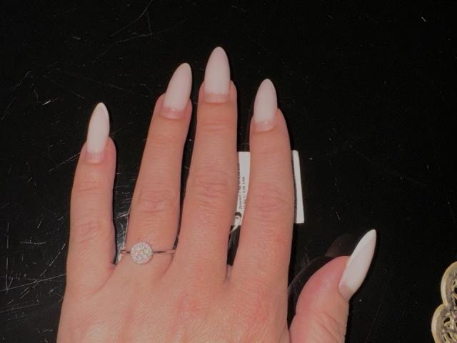 A mão de MysteryLaddy, com unhas longas, brancas e pontudas, e um anel com uma pequena pedra redonda, semelhante a um diamante, no dedo médio. Superfície preta, brilhante e texturizada abaixo..