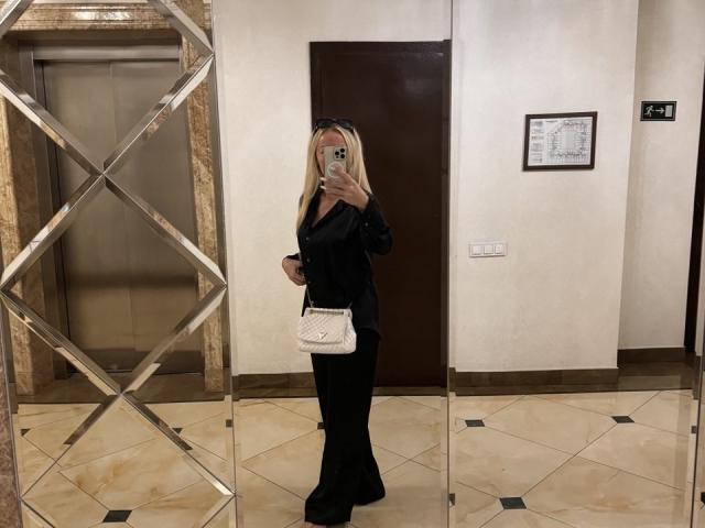MysteryLaddy with long blonde hair, black dress, and white quilted purse, taking a mirror selfie, showing her ass in the reflection. She's holding a phone with a green case.
