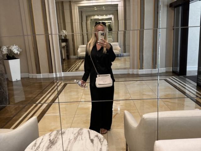 MysteryLaddy, debout devant un miroir, portant un haut noir à manches longues et une jupe noire, tenant un téléphone et un petit sac rose, avec des seins visibles..