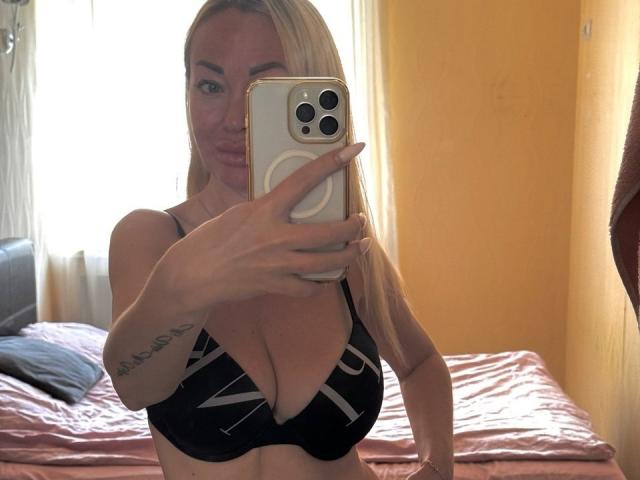 MysteryLaddy con cabello rubio, tomando una selfie en un espejo, mostrando sus grandes senos en un sostén negro, sosteniendo un teléfono blanco, tatuaje en el brazo derecho, expresión de labios fruncidos..