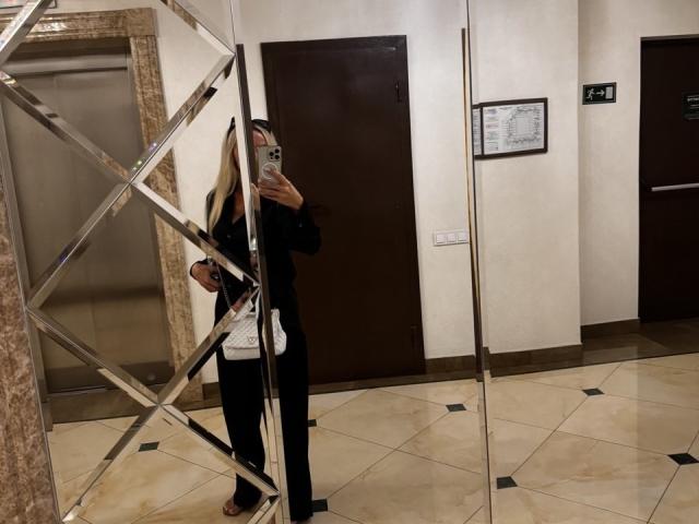 Une personne, MysteryLaddy, avec de longs cheveux blonds, vêtue de noir, prend un selfie devant un miroir dans une pièce carrelée, montrant le reflet de son visage et de son téléphone. Le miroir est situé à gauche..
