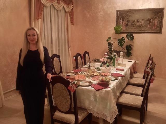 Een mysterieuze vrouw met lang blond haar, gekleed in een zwarte jurk, staat bij een eettafel met een witte tafelloper van kant, rode servetten en borden met eten, in een kamer met warme verlichting..