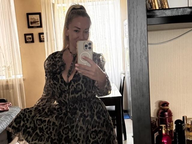 MysteryLaddy avec une queue de cheval blonde, portant une robe à imprimé léopard, tenant un téléphone pour un selfie devant un miroir, montrant sa poitrine, assise dans une pièce avec une table et une bibliothèque. Seins visibles..