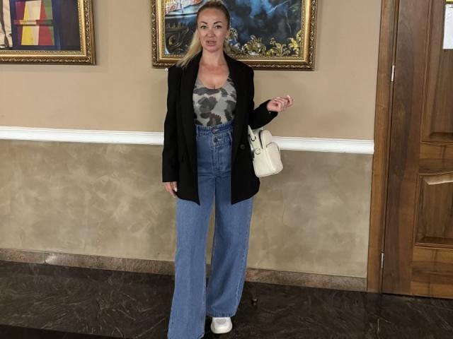 MysteryLaddy with blonde hair, black blazer, camo tank top, high-waisted blue jeans, white sneakers, holding white purse, standing in front of wooden door, marble floor.