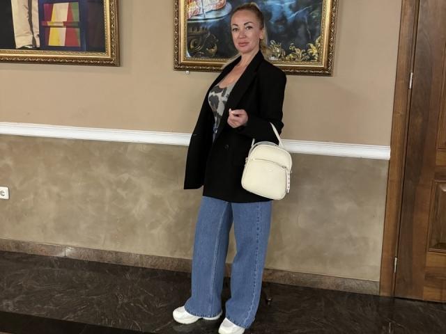 MysteryLaddy has blonde hair, white sneakers, blue flared jeans, black blazer, white handbag, and a gray top with a visible cleavage, standing in a room with paintings.