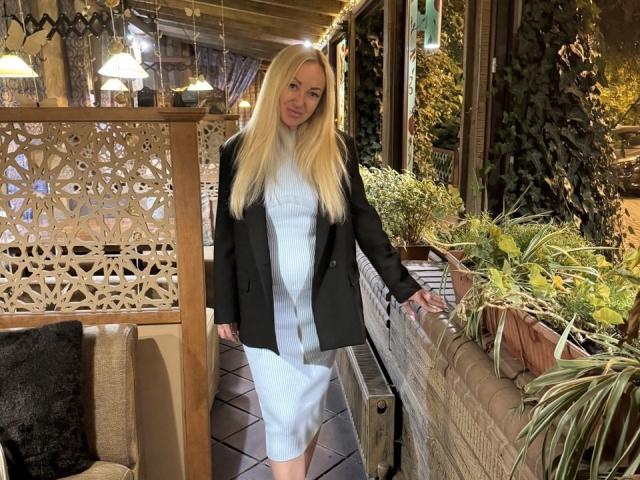 MysteryLaddy has long blonde hair, white dress, and black jacket, standing with a slight smile, holding a wooden wall, with potted plants and a cozy, lit interior.
