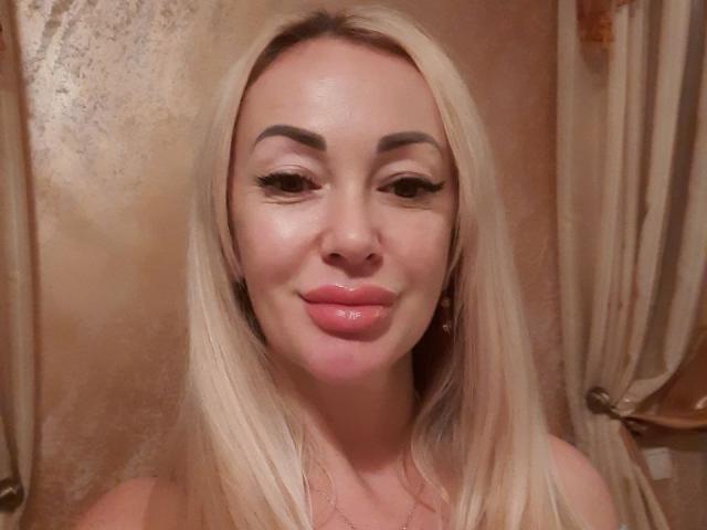 MysteryLaddy has shiny blonde hair, thick eyebrows, long eyelashes, and plump pink lips. She's smiling softly with a slight pinkish glow on her face.