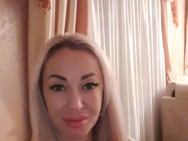 Een vrouw met lang blond haar, dikke wenkbrauwen en roze lippen, die glimlachend naar de camera kijkt en haar gezicht en bovenborst laat zien. Haar borsten zijn gedeeltelijk zichtbaar..