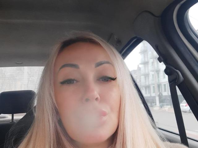 Una ragazza misteriosa con capelli biondi, sopracciglia folte e ciglia lunghe, labbra piene in un selfie leggermente sfocato, seduta in macchina, che guarda in avanti. Il seno non è visibile..