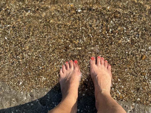 Stefanni's bare feet with red-painted toenails standing on a rocky, wet beach. Her ankles are visible, and she's slightly tanned. Her toes are slightly spread apart.