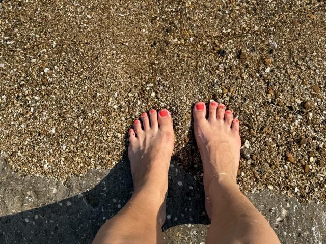 Stefanni's feet with bright red painted toenails standing on a sandy, rocky beach, visible from a top-down perspective. Her ankles are slightly bent, showing a small anklet.