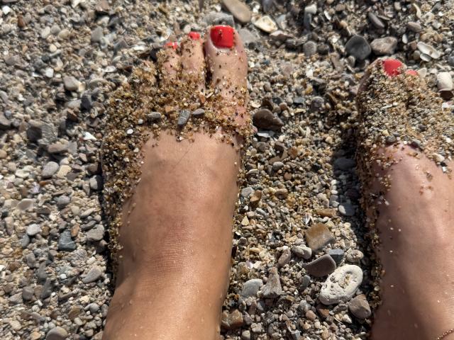 Stefanni's feet with red-painted toenails, covered in sandy pebbles, standing on a rocky beach. Sand and small rocks cling to her toes and lower legs.
