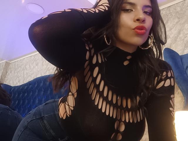 VioletaStone with dark hair, pouty red lips, wearing a black, cut-out top, and blue jeans, leaning forward, hand in hair, showing her round, exposed ass.