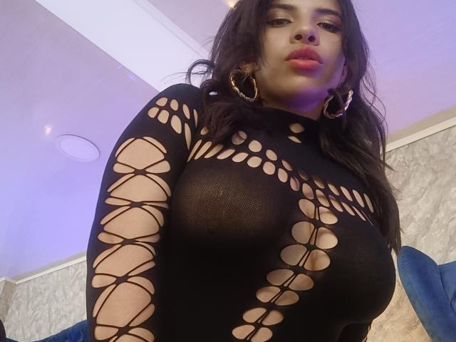 VioletaStone in a black, see-through, lace-up top, her big boobs clearly visible through the holes, wearing large gold hoop earrings, with wavy dark hair and pink lipstick.