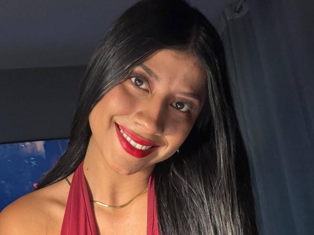 MadinssonGrey has long black hair, red lipstick, and a gold necklace, smiling with red halter top, showing her shiny tits and nose ring.