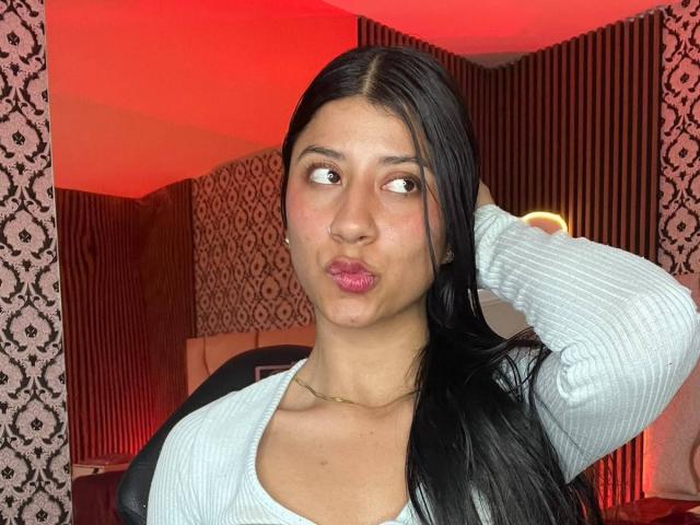MaddinssonGrey with pouty lips, black hair, and light blue ribbed shirt, hand in hair, looking sideways, red lighting, patterned wallpaper, and a ring light in the background.