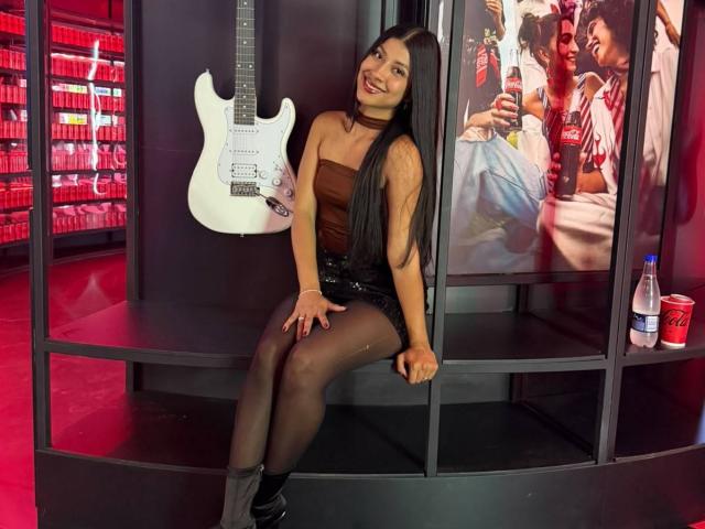 MadinssonGrey con cabello negro largo, un top marrón sin tirantes, pantalones cortos negros con lentejuelas, medias negras y botas grises, sentada con una guitarra blanca, sonriendo, fondo de color rojo, una botella de refresco a la derecha..