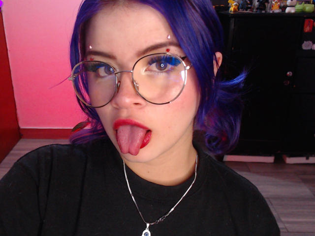 RavenThuwu with purple hair, glasses, red lipstick, and tongue out, wearing a black shirt and silver necklace, in a selfie. Boobless, no visible ass or pussy.