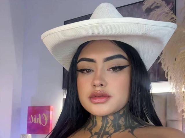 LairaHearvy with black hair, white cowboy hat, tattooed neck, thick eyebrows, winged eyeliner, and pink glossy lips, looking at camera. Her ass is not visible.