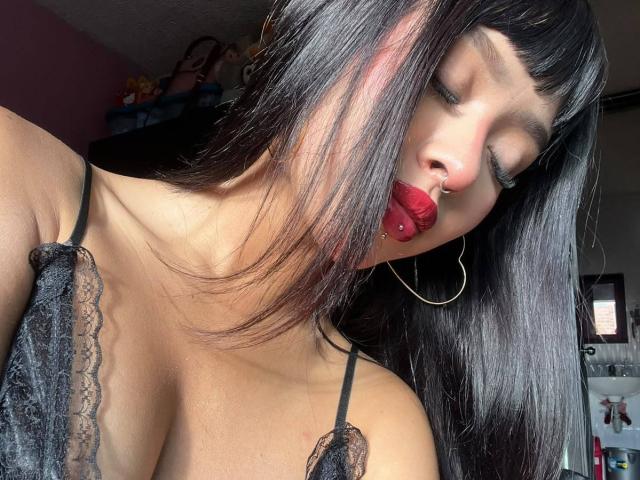 AdaraVance, avec ses cheveux noirs et lisses, son rouge à lèvres rouge, son anneau de nez et son boucle d'oreille, porte un haut en dentelle noire qui révèle ses seins, et a un piercing sur la lèvre, dans une pièce ensoleillée..