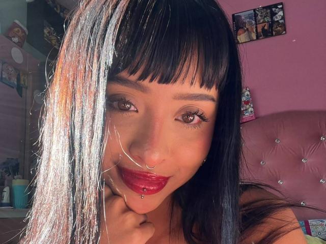 AdaraVance met zwart haar en witte highlights, bruine ogen, rode lipstick, een neusring en een doorboorde onderlip, lachend met zonlicht op haar gezicht, in een roze slaapkamer..