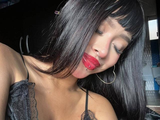 AdaraVance, con i capelli neri, rossetto rosso, piercing al naso e orecchini a cerchio, occhi chiusi, indossa una camicetta di pizzo nera e si sporge leggermente in avanti..