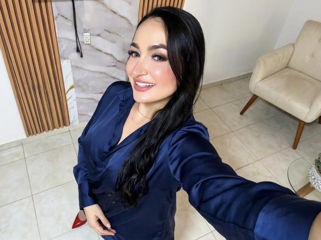 PamelaLiss with long black hair, wearing a navy blue satin shirt, smiling, showing red pointed nails, taking a selfie, holding out her arm, tits visible under shirt.