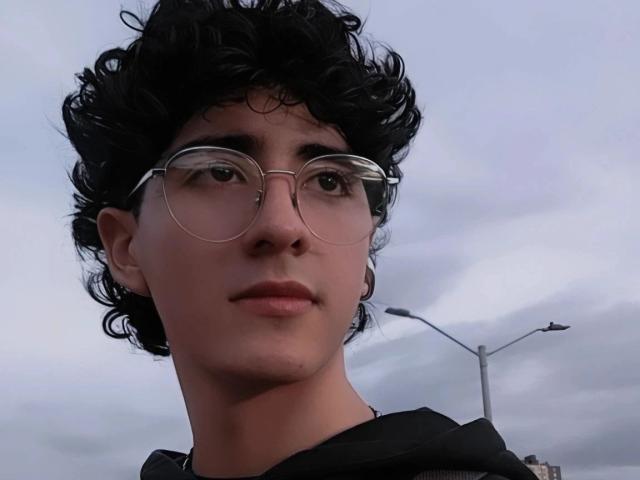 ElliotNoir has curly black hair, wearing round glasses, with a slight smile, and a black hoodie. His skin is tan, and he's looking up with a relaxed expression.