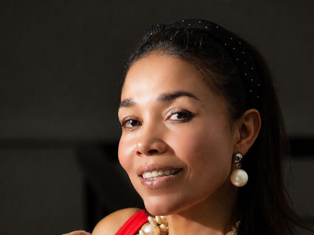 MiaLucchi has brown skin, dark eyes, and a cute smile. She's wearing a red top, pearl earrings, and a black headband with small white dots. Her tits are small.