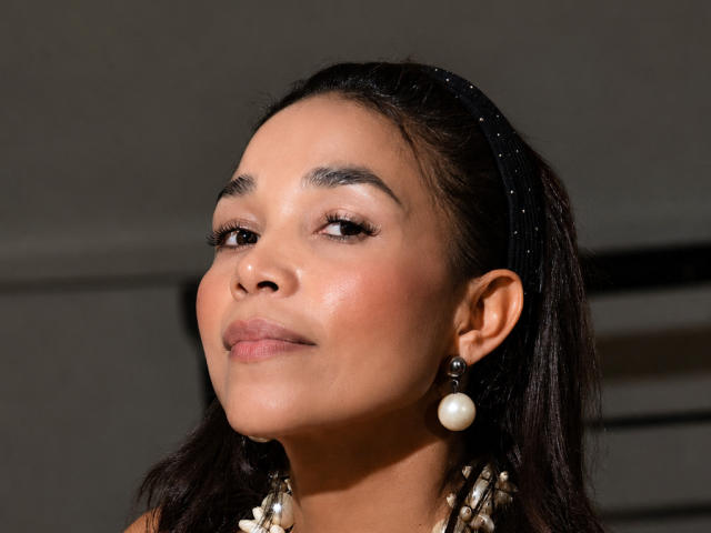 MiaLucchi with medium brown skin, dark hair in a headband, wearing pearl earrings, and a necklace, has a subtle smile, and soft, shiny makeup on her face.