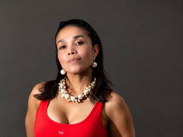 MiaLucchi with medium tits in a red tank top, wearing a pearl necklace and earrings, dark hair, slight smile, gray background, looking at camera.