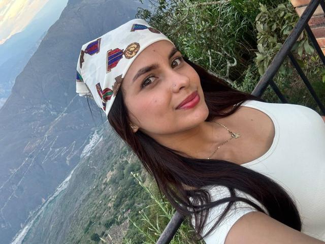 DulceColeman with long dark hair, white headscarf, colorful patterns, white top, red lipstick, small nose ring, and necklace, showing her tits in a mountain selfie.