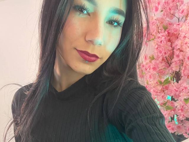 VioletaVelvet with long black hair, dark lipstick, and greenish eye makeup, wearing a black ribbed sweater, standing next to pink cherry blossoms.