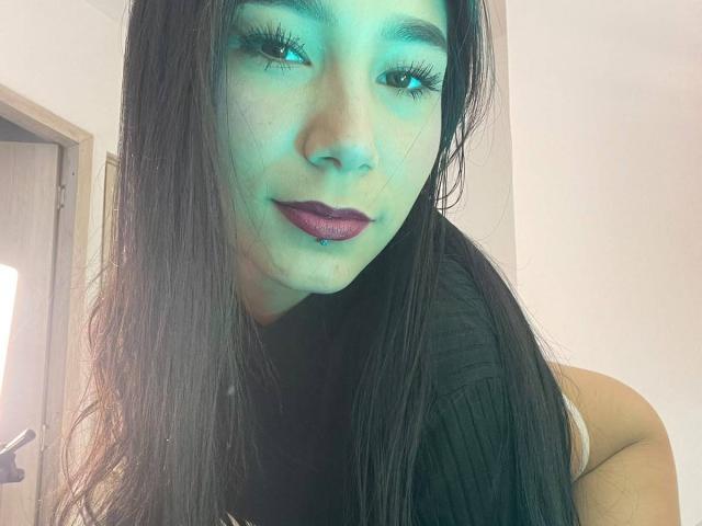 VioletaVelvet with long dark hair, green-tinted face, purple lipstick, and a lip piercing, looking at the camera with a slight smile, wearing a black top.