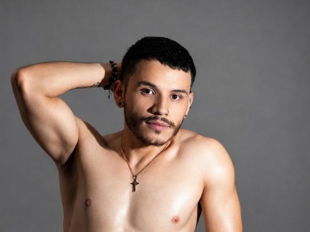 LianFerrer, shirtless, with a cross necklace, short black hair, and a beard, raising his right arm, showing a muscular, tan, and sweaty chest. Gray background.