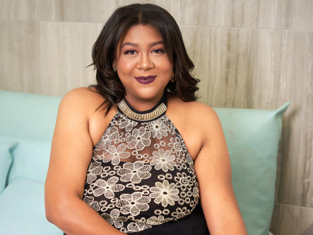 BiancaForest with dark wavy hair, brown skin, and purple lipstick, wearing a black halter top with gold floral lace, showing her tits and ass, sitting on a green pillow.