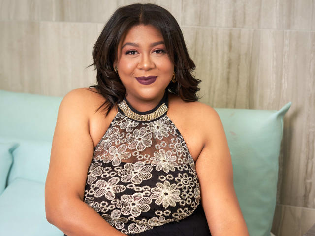BiancaForest with medium brown skin, dark wavy hair, wearing a black halter top with gold floral embroidery, dark purple lipstick, gold earrings, sitting on a light blue pillow.