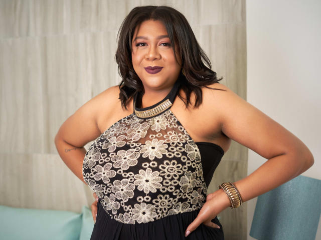 BiancaForest, a curvy woman with dark brown hair, wearing a black halter dress with silver floral lace, has her hands on her hips, smiling confidently.