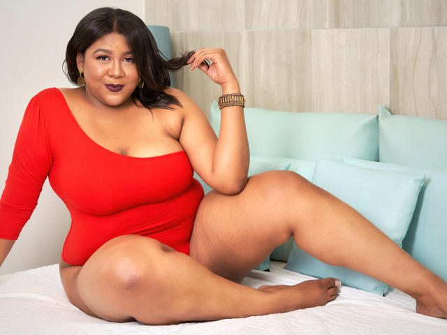 BiancaForest, une femme corpulente aux cheveux foncés, portant une combinaison rouge à une épaule, assise sur un lit, touchant ses cheveux, montrant ses seins et ses parties intimes, souriant..