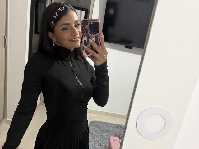 KimmyK avec des cheveux foncés, des barrettes en forme d'étoile rose, une robe noire à fermeture éclair, tenant un téléphone rose, souriante, selfie devant un miroir, chaussures plates noires, bague sur le mur, mur blanc, sol carrelé, reflet dans le miroir..