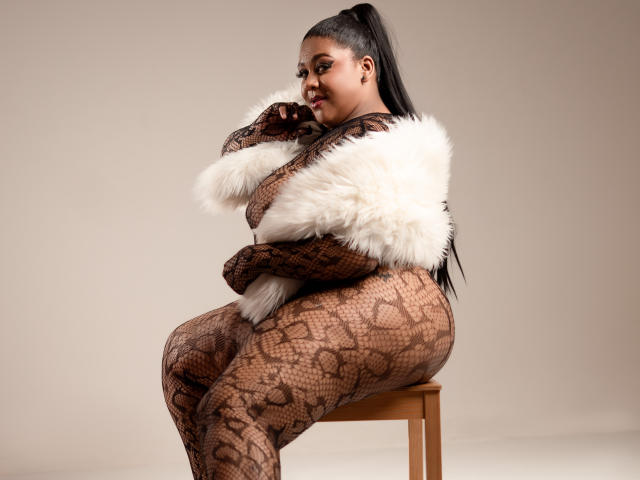 BiancaForest, curvy with large ass, wearing snake-patterned fishnet bodysuit, white fur shawl, sitting on wooden stool, smiling, black ponytail, brown skin, looking over shoulder.