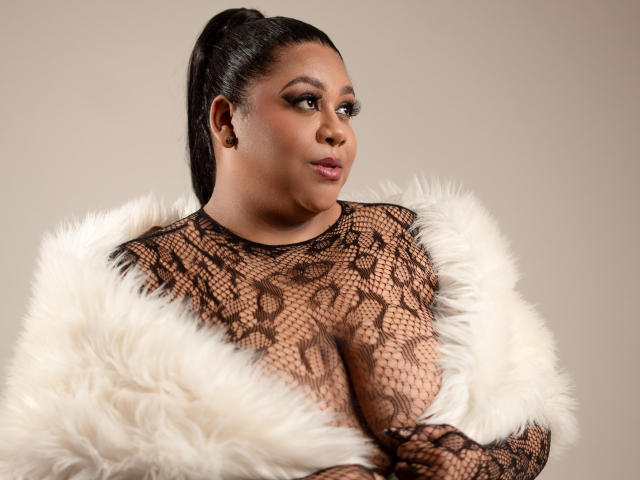 BiancaForest with large tits, wearing black lace top, white fur wrap, dark skin, long black hair in ponytail, looking to the side, neutral expression.