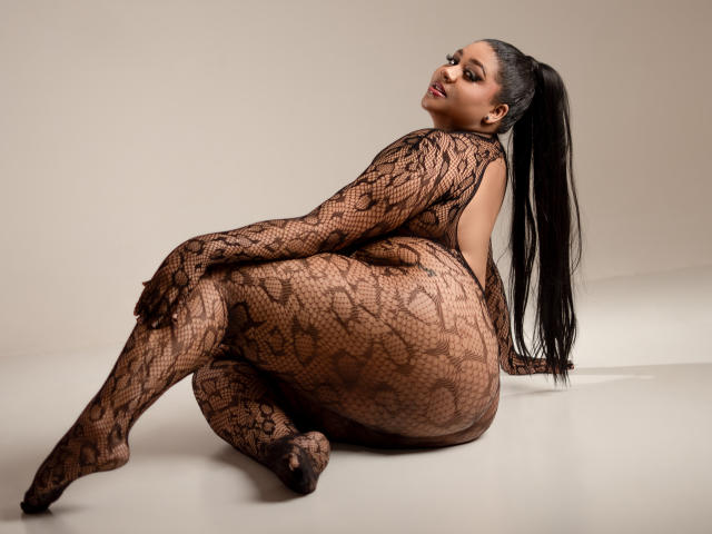 BiancaForest with long black hair in a ponytail, wearing a black lace bodysuit, sitting on the floor, looking back with a seductive smile, her ass and pussy visible.